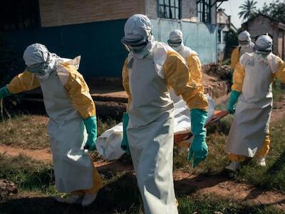 Miembros del personal de MSF, equipados con material de protección individual, trasladan el cuerpo de un paciente fallecido en el Centro de Tratamiento del Ébola a la nueva morgue habilitada dentro del Centro de Tratamiento del Ébola gestionado por MSF en el Hospital General de Mongbwalu. La nueva morgue permite a las familias ver a sus seres queridos antes de que los cuerpos sean depositados en ataúdes. Mogwalu, Ituri, 21 de junio de 2026. © Alexis Huguet/MSF