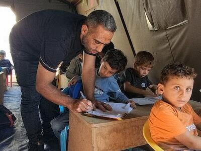 Noticias ONU Un maestro ayuda a niños palestinos en un aula instalada en una tienda de campaña.