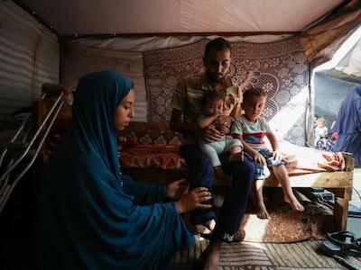 Bassel sostiene a sus hijos, Mohammed, de nueve meses, e Ibrahim, de dos años y medio, mientras su esposa le masajea la pierna amputada para aliviar el dolor fantasma. La familia vive en una tienda de campaña para desplazados en Mawasi, Jan Yunis, tras huir de su hogar en Rafah. Bassel perdió la pierna tras recibir un disparo meses antes cuando intentaba acceder a la ayuda en un punto de distribución. © Motasem Abu Aser/MSF