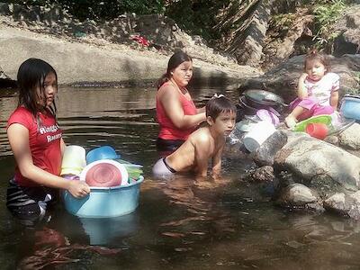 - Una madre lava utensilios de cocina en una poza del río Aguas Calientes, mientras sus hijos se divierten. Ella contó a IPS que ese riachuelo, parte de la cuenca del río Lempa, el más largo de El Salvador, siempre tenía un abundante caudal, pero ahora por el cambio climático y el uso de agua para riego de caña de azúcar su nivel ha disminuido. Foto por Edgardo Ayala/IPS